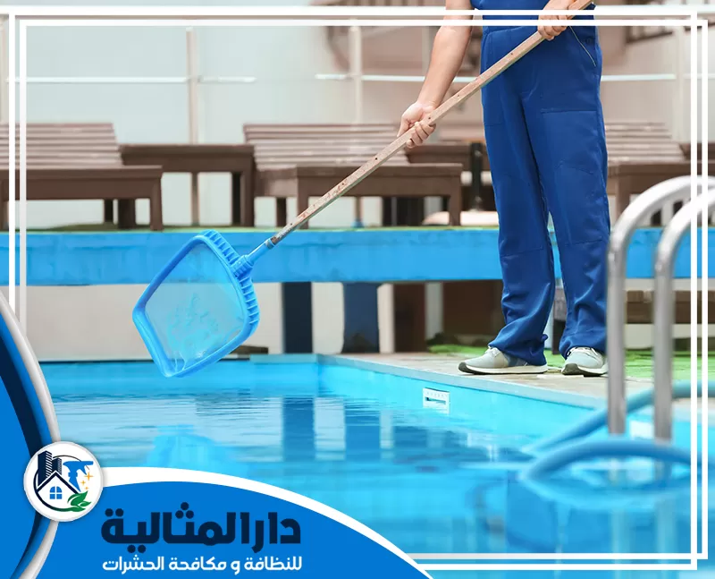 شركة تنظيف مسابح بالقطيف | 0507082455 | دار المثالية رجل يرتدي زيًا أزرق يحمل ممسحة أمام مسبح، يُمثل خدمة تنظيف مسابح في القطيف.